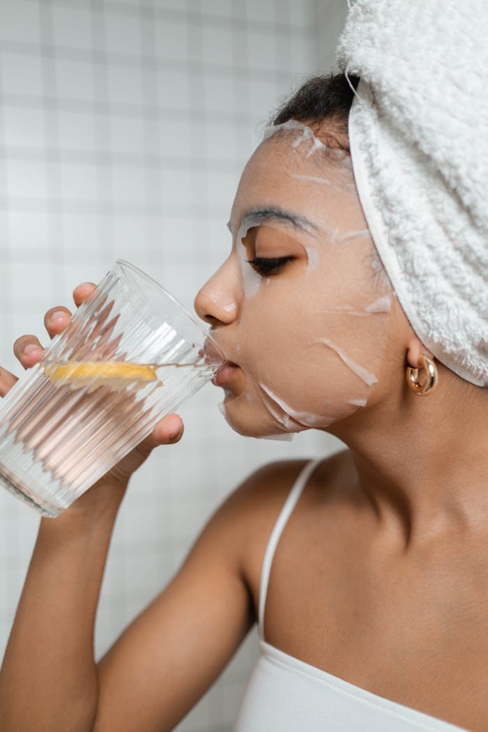 Woman in skincare routine with sheet mask, drinking lemon water for hydration.